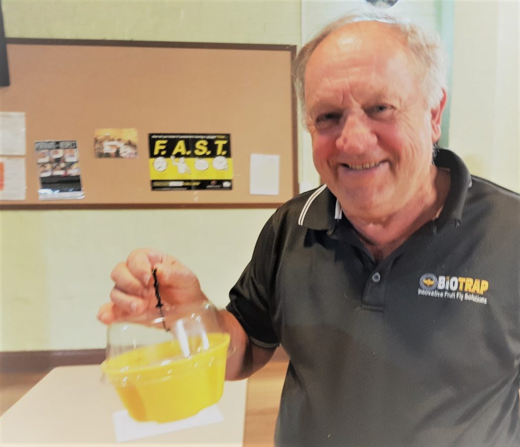 Fruit fly trap inventor Colin Bain looking at the camera wearing a tshirt branded "Biotrap: Innovative Fruit Fly Services". He's holding a Biotrap and smiling at the camera.