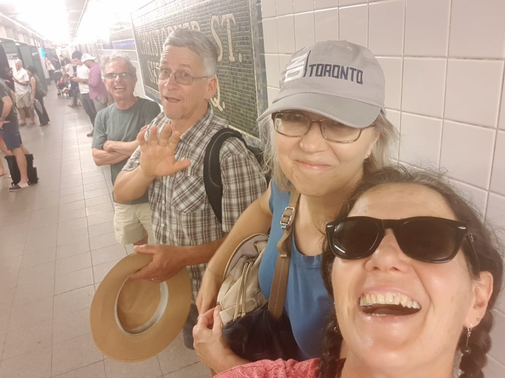 Two men and two women standing against a wall in an underground railway station in front of a sign that says Christopher St. Everyone is smiling and one of the men is waving.