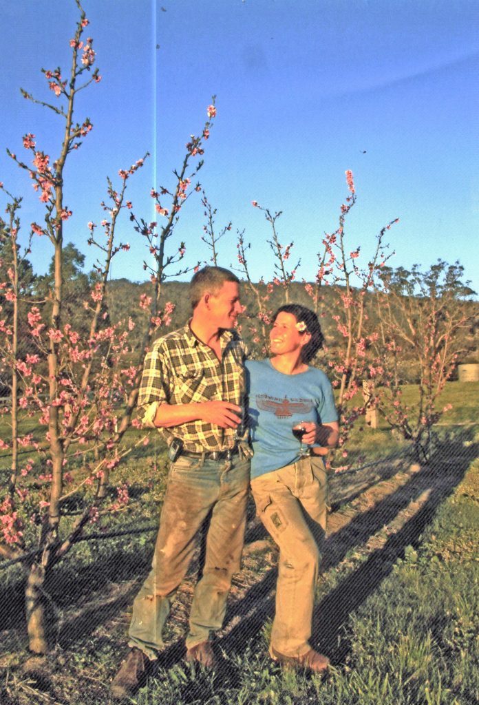 A man in his forties in a checked shirt and jeans and a woman wearing a blue tshirt and work jeans standing in front of a row of peach trees with pink flowers. The setting afternoon sun is on their faces as they smile towards each other, both holding a glass of wine.