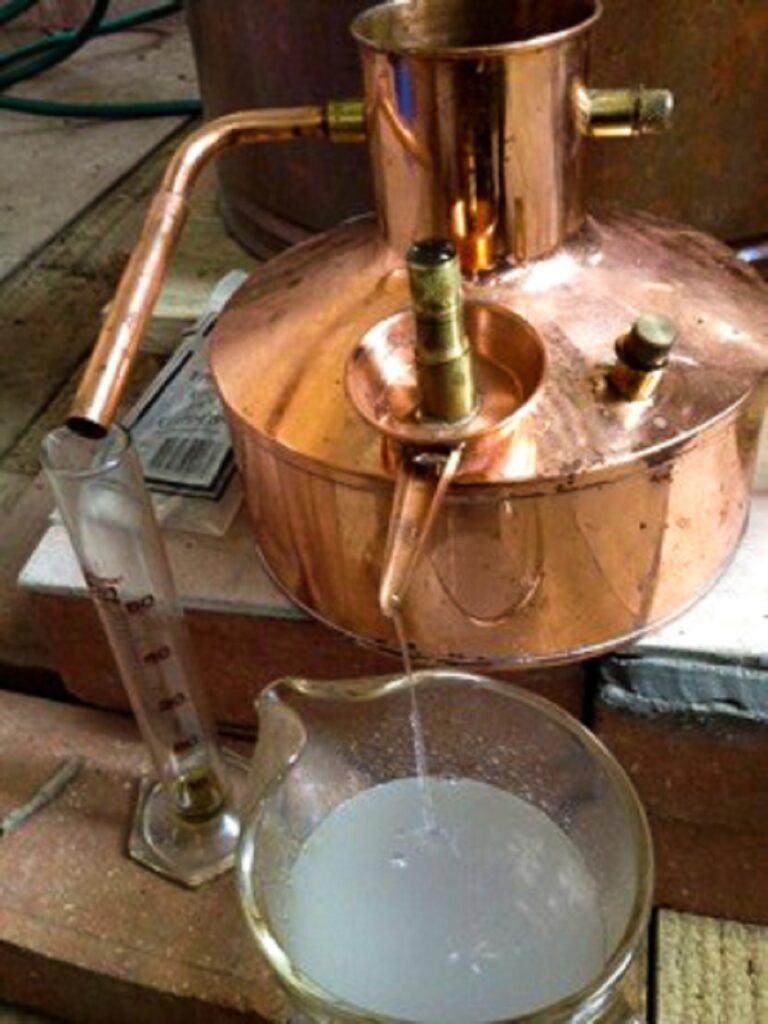 Liquid neroli hydrosol draining from a copper vessel while neroli essential oil drips into a test tube nearby.