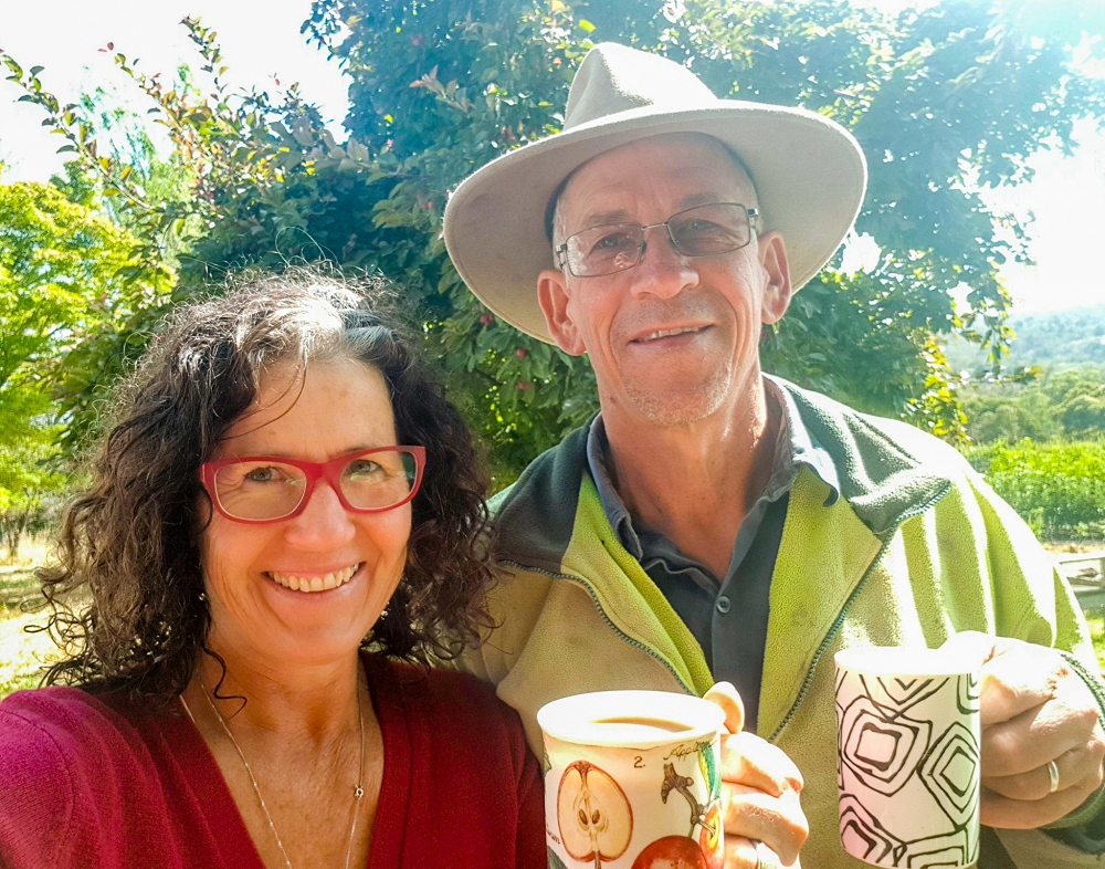Katie Hugh coffee garden-1000x786 A woman in a red cardigan and a man wearing a hat and geen jumper holding coffee cups in the garden