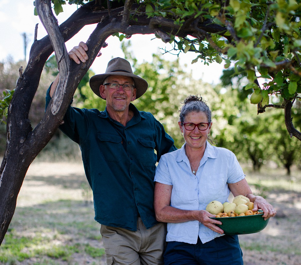 Katie Hugh green bowl fruit square-1000x881 A man in a hat and green shirt leaning on a tree and standing next to a woman in a short-sleeve blue shirt holding a green bowl full of fruit.