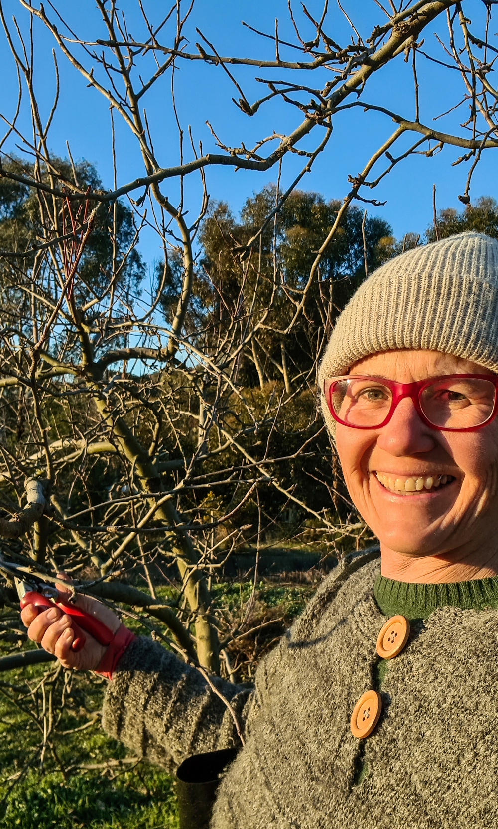 Katie pruning woolly jumper-1000x1662 A woman wearing a grey beanie and woolly jumper smiling at the camera while pruning a dormant fruit tree